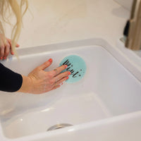 A hand using a blue "Mint" sponge to clean a white sink, showcasing the product's functionality in a home setting.