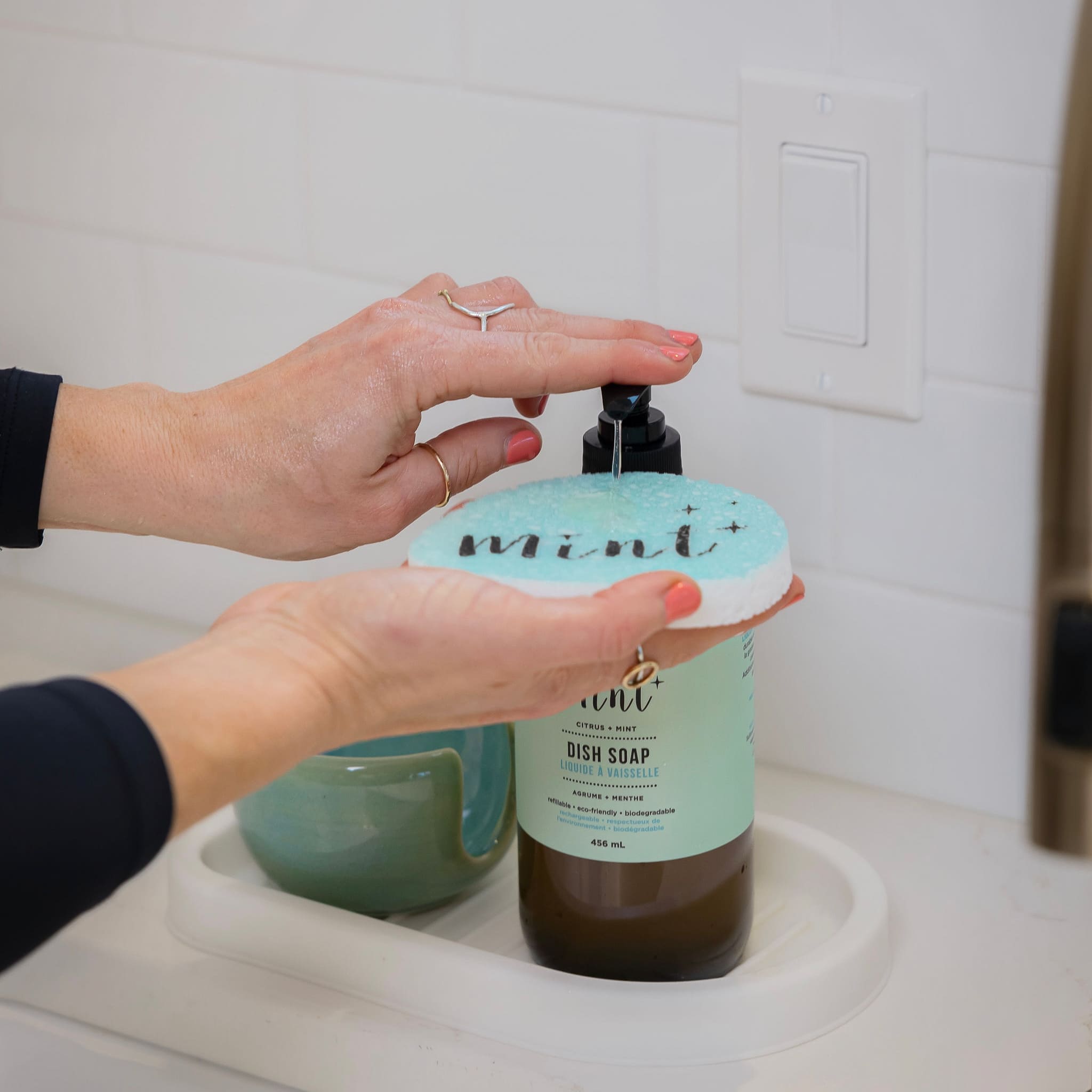 A hand applying dish soap onto the &quot;Mint&quot; sponge, with a pump bottle in the background, illustrating the sponge’s use in dish cleaning.