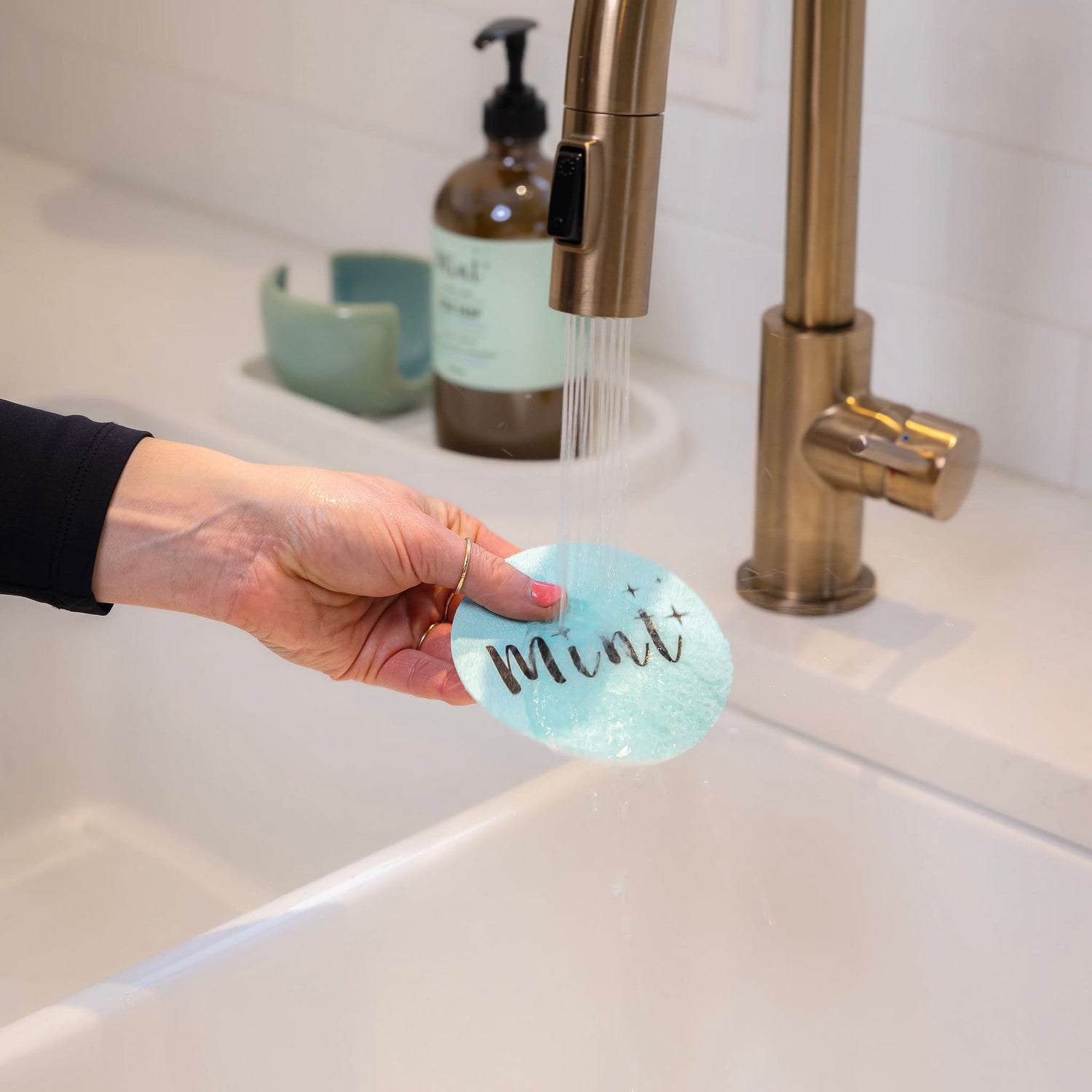 A hand rinsing the &quot;Mint&quot; sponge under a kitchen faucet, with water running over it, emphasizing the sponge&