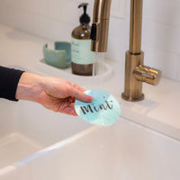 A hand rinsing the "Mint" sponge under a kitchen faucet, with water running over it, emphasizing the sponge's easy-to-clean design.