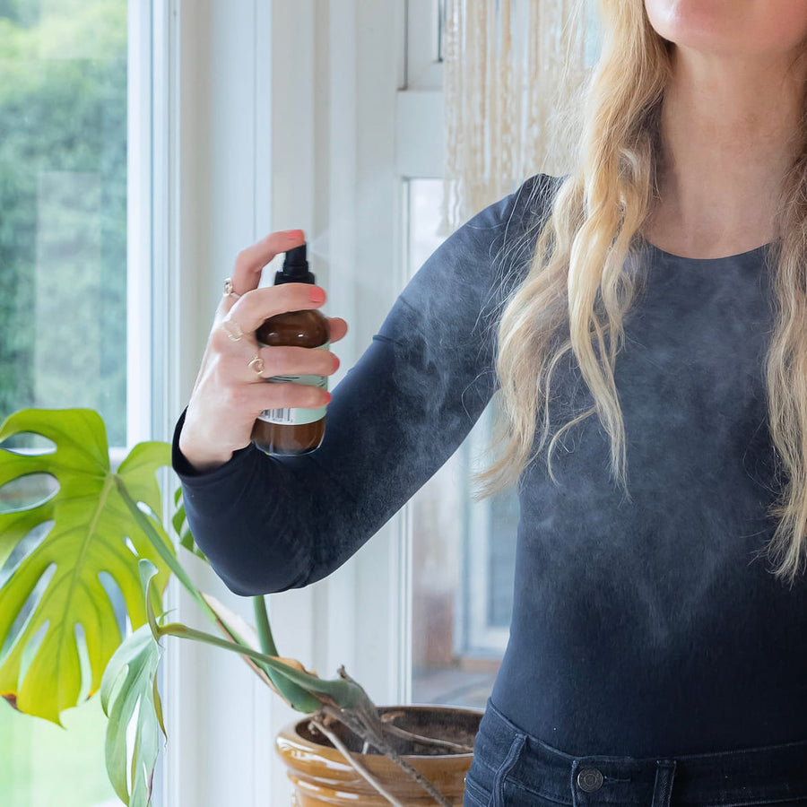 Person using Mint Winter Mist Linen + Room Spray, spraying mist in the air in front of a leafy green houseplant.