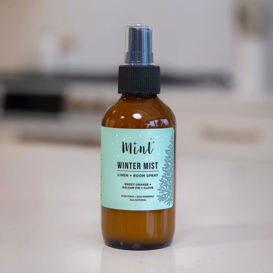 Close-up of Mint Winter Mist Linen + Room Spray bottle on a kitchen counter, showcasing its eco-friendly label and amber glass packaging