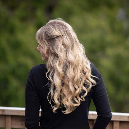 The image shows a person with long, wavy blonde hair styled with soft curls, viewed from the back. They are wearing a black top and standing outdoors on a wooden deck, with a blurred green background suggesting a natural setting.