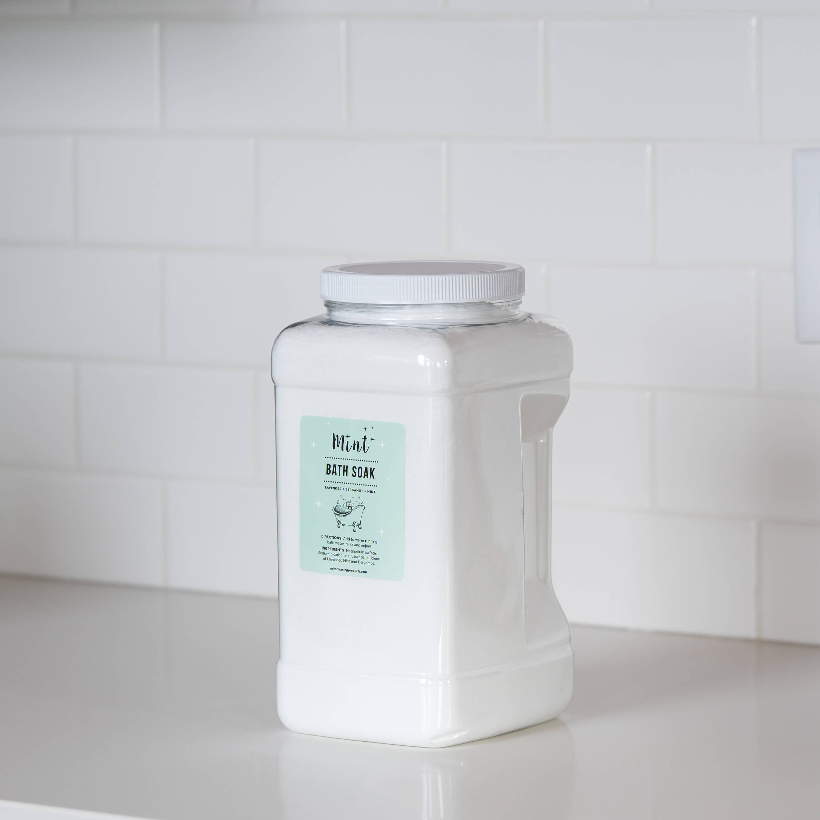 Bath Soak: A large white plastic container of &quot;Mint+ Bath Soak&quot; displayed on a white countertop with a clean subway tile backsplash. The mint green label highlights the calming and natural properties of the product.