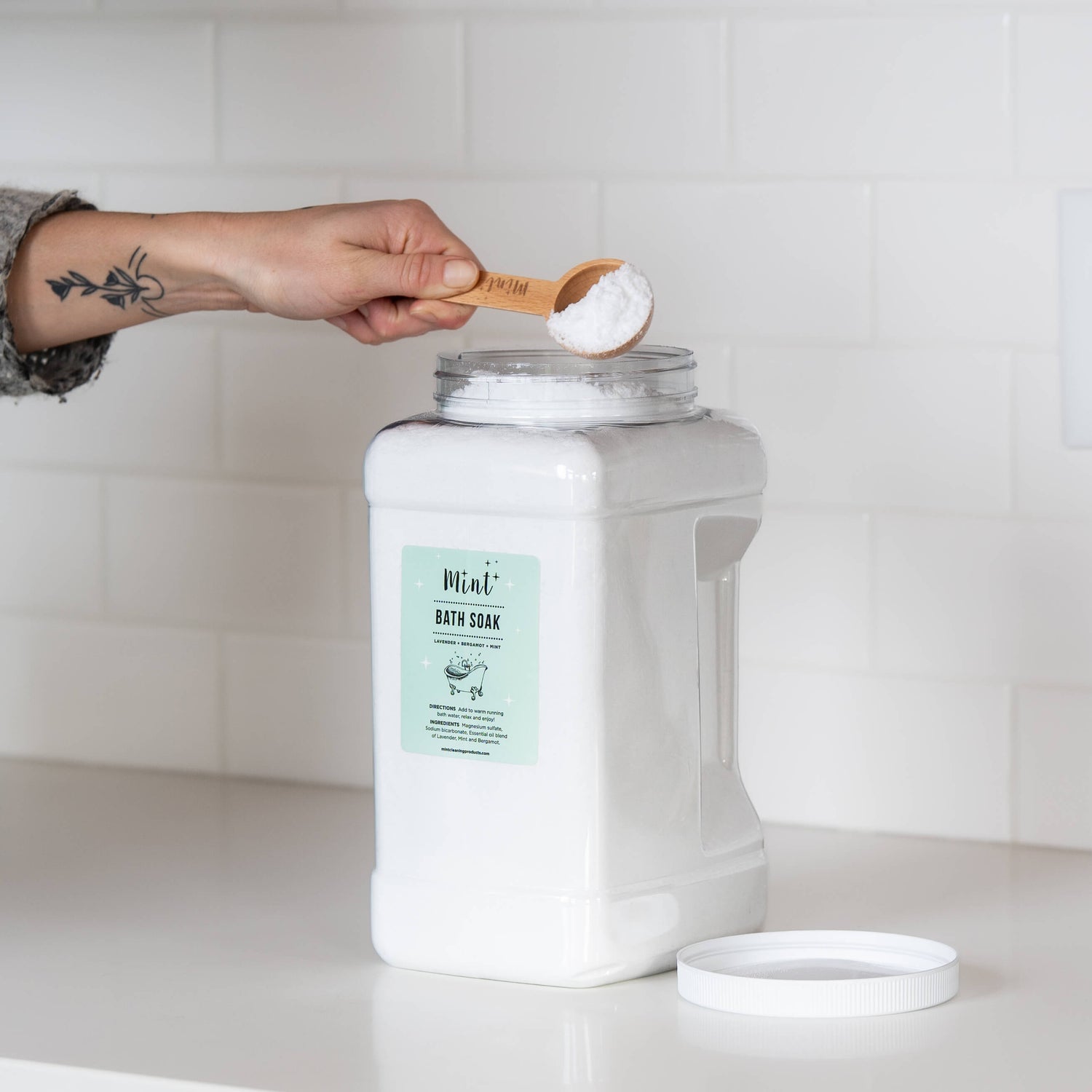 A large white container of &quot;Mint+ Bath Soak&quot; on a clean white countertop with its lid removed. A hand holding a bamboo scoop filled with the product is positioned above the container, set against a subway tile backsplash.