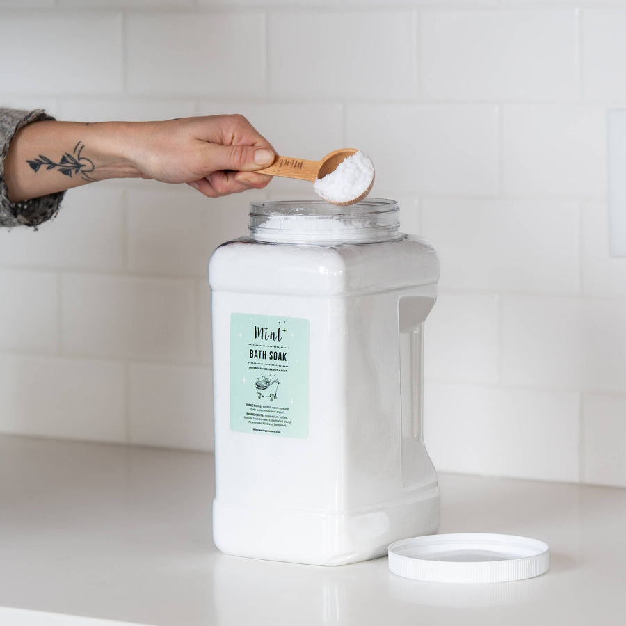 A large white container of "Mint+ Bath Soak" on a clean white countertop with its lid removed. A hand holding a bamboo scoop filled with the product is positioned above the container, set against a subway tile backsplash.