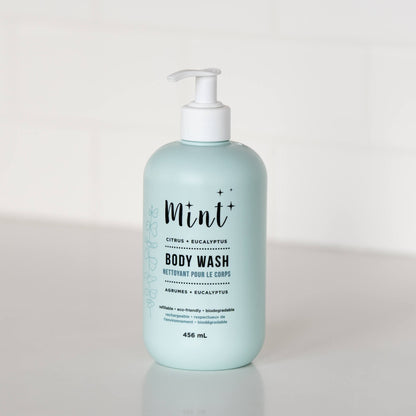 A 456 mL light blue bottle of &quot;Mint+ Body Wash&quot; with a pump dispenser, displayed on a white countertop. The label features the scents &quot;Citrus + Eucalyptus&quot; with bilingual text &quot;Nettoyant pour le corps&quot; and highlights eco-friendly attributes such as being refillable, biodegradable, and sustainable. Subtle botanical illustrations adorn the left side of the label, creating a clean and natural presentation. The background includes a soft, minimalistic white setting.