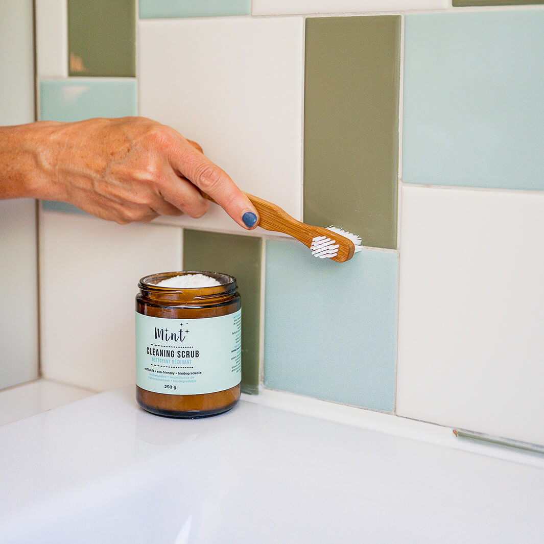 Hand using a bamboo brush to clean grout on green and white tiled wall, with a jar of Mint Cleaning Scrub labeled &