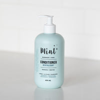 A 456 mL light blue bottle of "Mint+ Conditioner" with a pump dispenser, displayed on a white countertop. The label highlights the scent "Rosemary + Mint" and includes bilingual text, "Revitalisant" and "Romarin + Menthe." It emphasizes eco-friendly attributes such as being refillable, biodegradable, and sustainable. A subtle botanical illustration adorns the left side of the label, adding a natural element to the design. The background features a bright and minimalistic white setting.