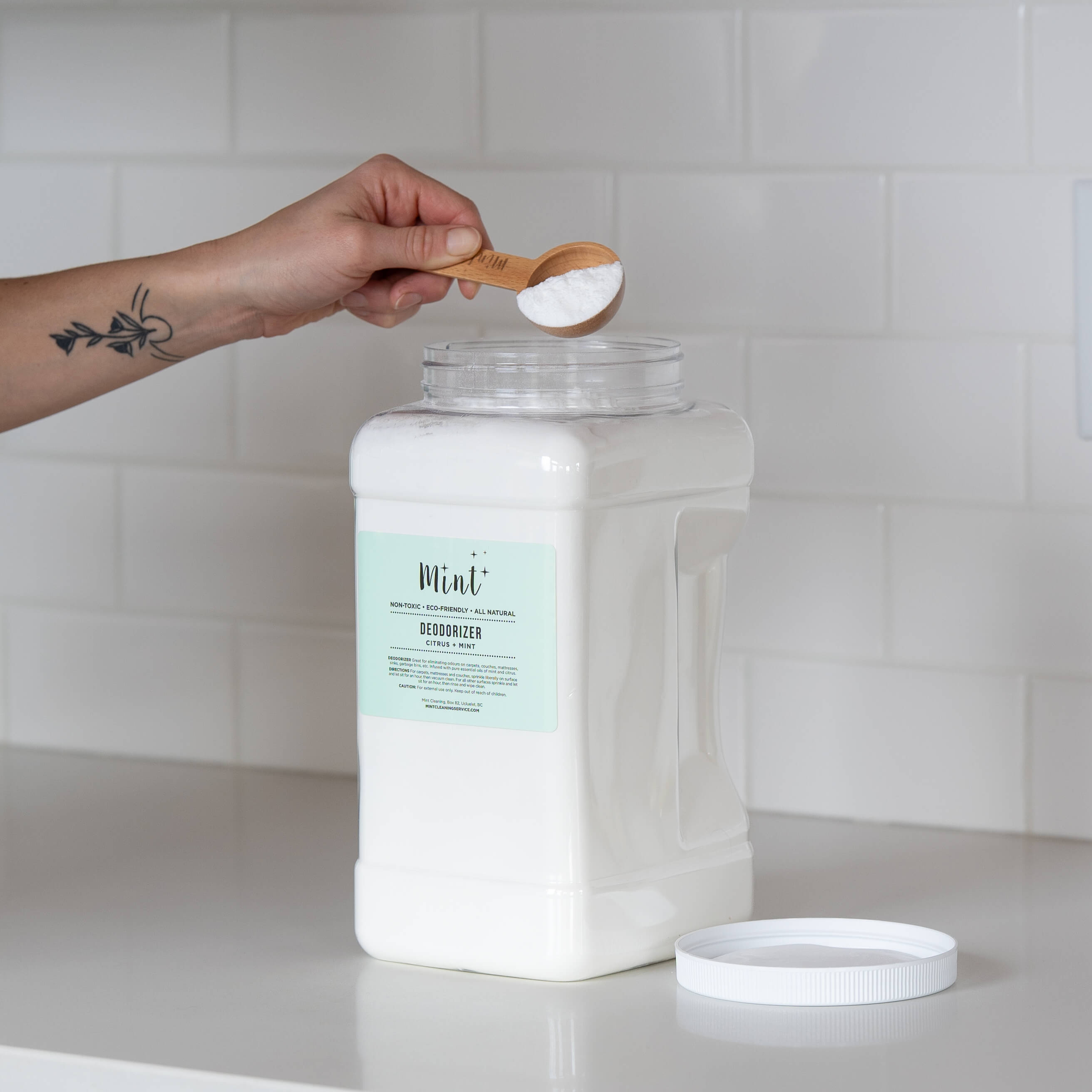 A large white container of &quot;Mint+ Deodorizer - Citrus + Mint&quot; with its lid off, set on a sleek white countertop. A hand with a bamboo scoop filled with the product is held above the container, against a bright subway tile backdrop.