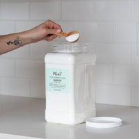 A large white container of "Mint+ Deodorizer - Citrus + Mint" with its lid off, set on a sleek white countertop. A hand with a bamboo scoop filled with the product is held above the container, against a bright subway tile backdrop.