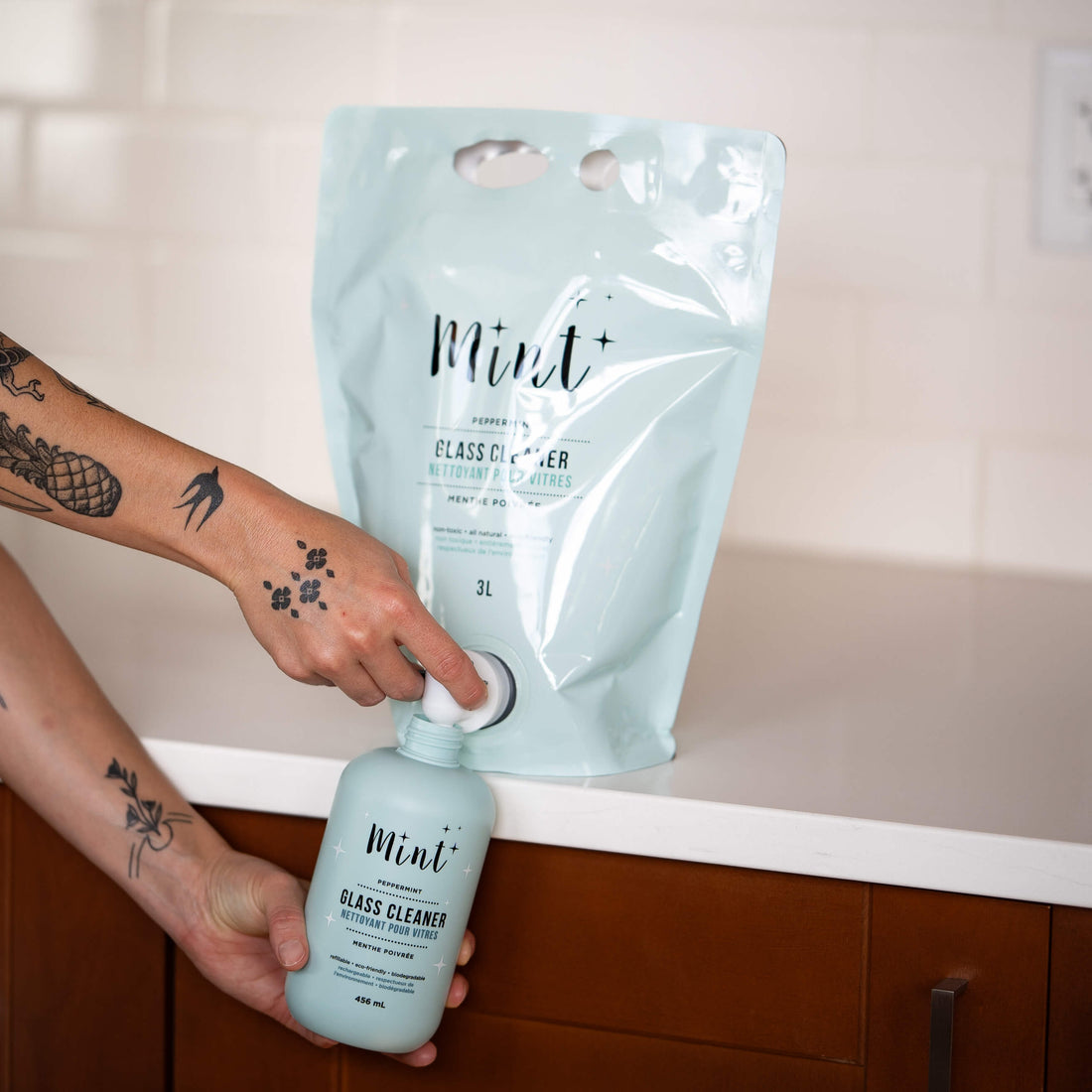 Hand with tattoos refilling a reusable light blue bottle labeled &