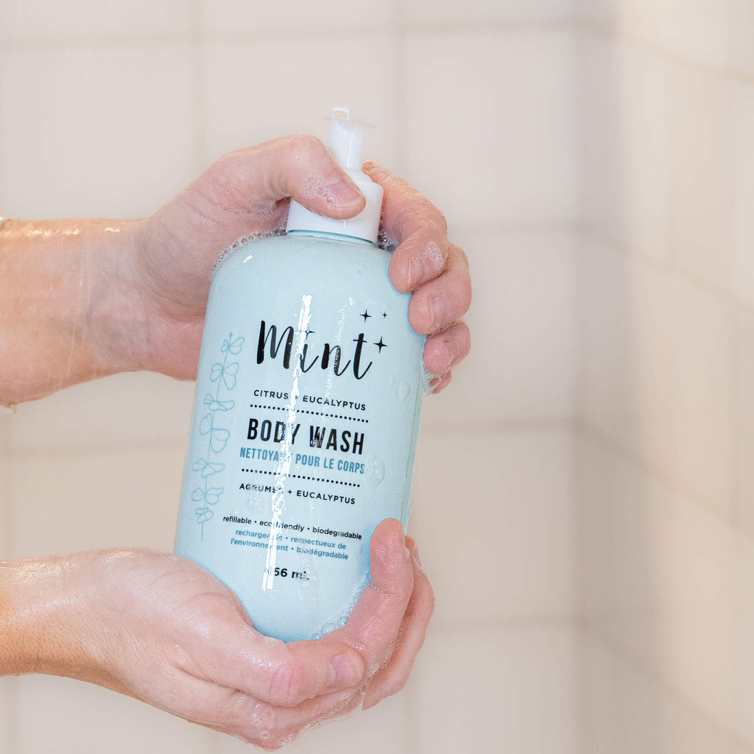 Hands holding a light blue bottle of Mint Citrus + Eucalyptus Body Wash in a tiled shower, with water and bubbles on the bottle.