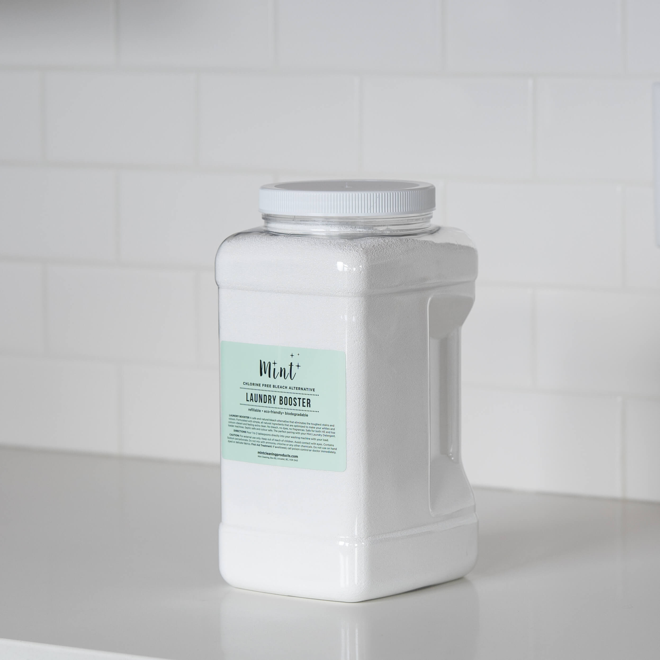 A large white plastic container of &quot;Mint+ Laundry Booster&quot; positioned on a modern white countertop with a neutral subway tile backsplash, highlighting its chlorine-free bleach alternative.