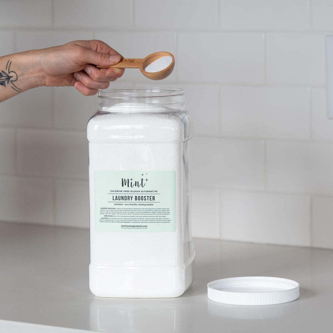 A large white container of &quot;Mint+ Laundry Booster&quot; displayed on a white countertop. The lid is off, and a tattooed hand is holding a bamboo scoop with product above the open container, with a clean subway tile backsplash in the background.