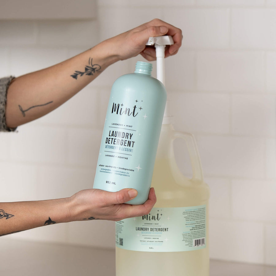 A person refilling a 952 mL light blue bottle of "Mint+ Laundry Detergent" with the scent "Lavender + Mint" from a large 3.8 L refill jug. The smaller bottle is labeled with bilingual text and eco-friendly features, such as being refillable and biodegradable. The person’s tattooed hand holds the pump on the large jug, with the smaller bottle positioned below. The background features a clean white countertop and tiled wall.