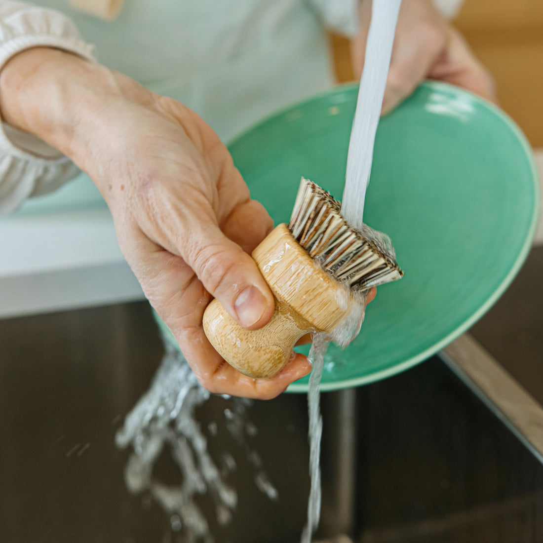 A person washing a green plate using a bamboo dish brush under running water, showcasing an eco-friendly cleaning tool in action.