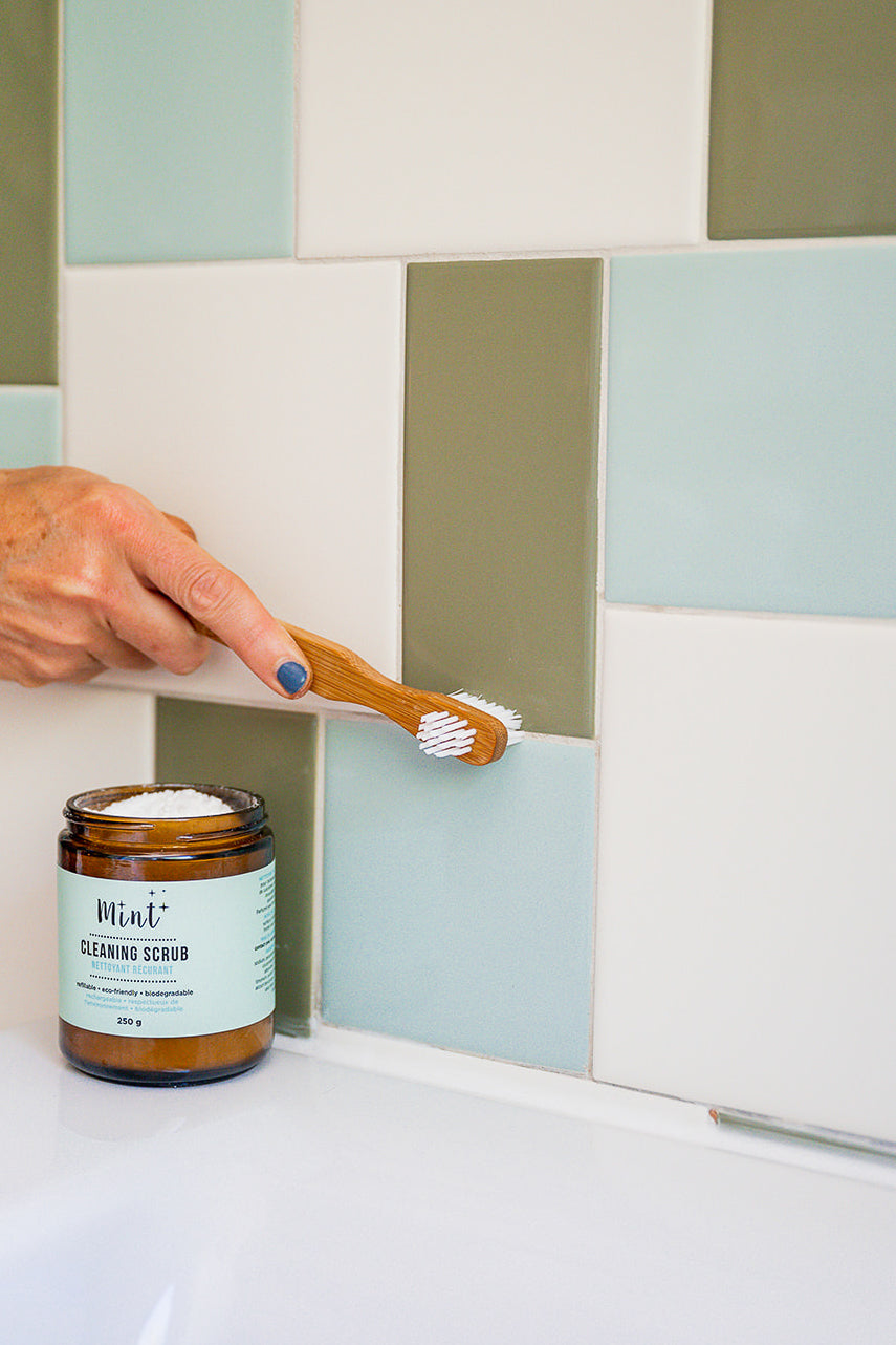 A person cleaning grout between colourful tiles using a Mint bamboo toothbrush and Mint Cleaning Scrub, displayed in an amber glass jar on a sink ledge.