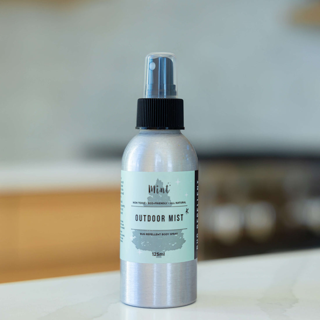 A 125ml aluminum bottle of Mint Outdoor Mist with a black spray top, labeled as a non-toxic and eco-friendly bug-repellent body spray, displayed on a countertop with a blurred background.