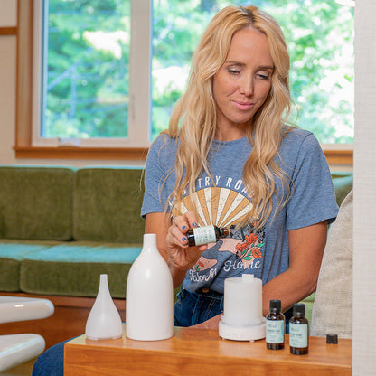 A woman sits by a diffuser, adding Mint essential oil from a glass bottle labeled &