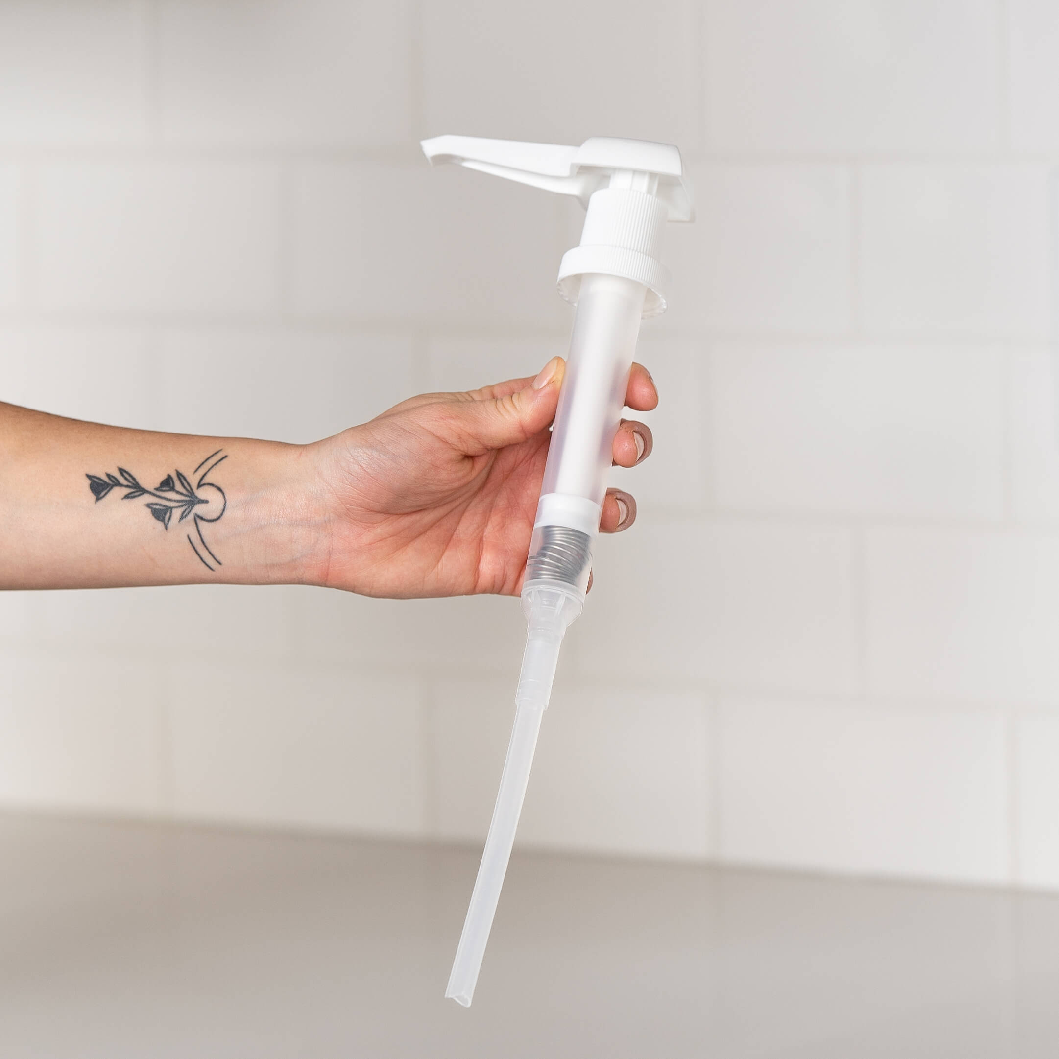 Close-up of a hand holding a white pump dispenser with a long tube, set against a clean, tiled background. The arm features a minimalist botanical tattoo, adding a personal touch to the image.