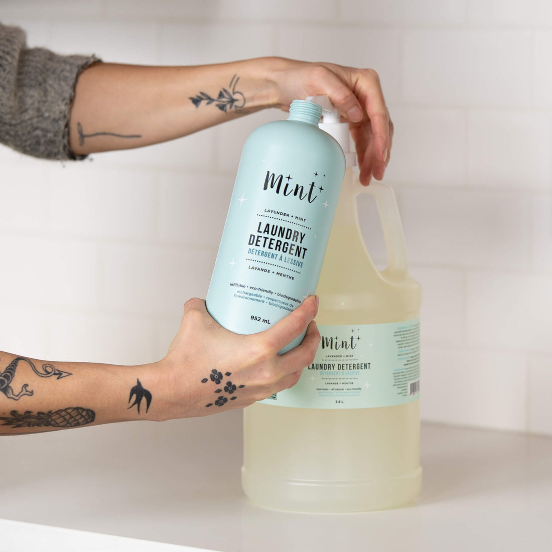 A person holding a 952 mL light blue bottle of "Mint+ Laundry Detergent" with the scent "Lavender + Mint," being refilled from a 3.8 L refill jug. The smaller bottle features bilingual text and eco-friendly attributes, such as being refillable and biodegradable. The person’s tattooed hands are visible, with the large jug in the background on a white countertop with a tiled wall.