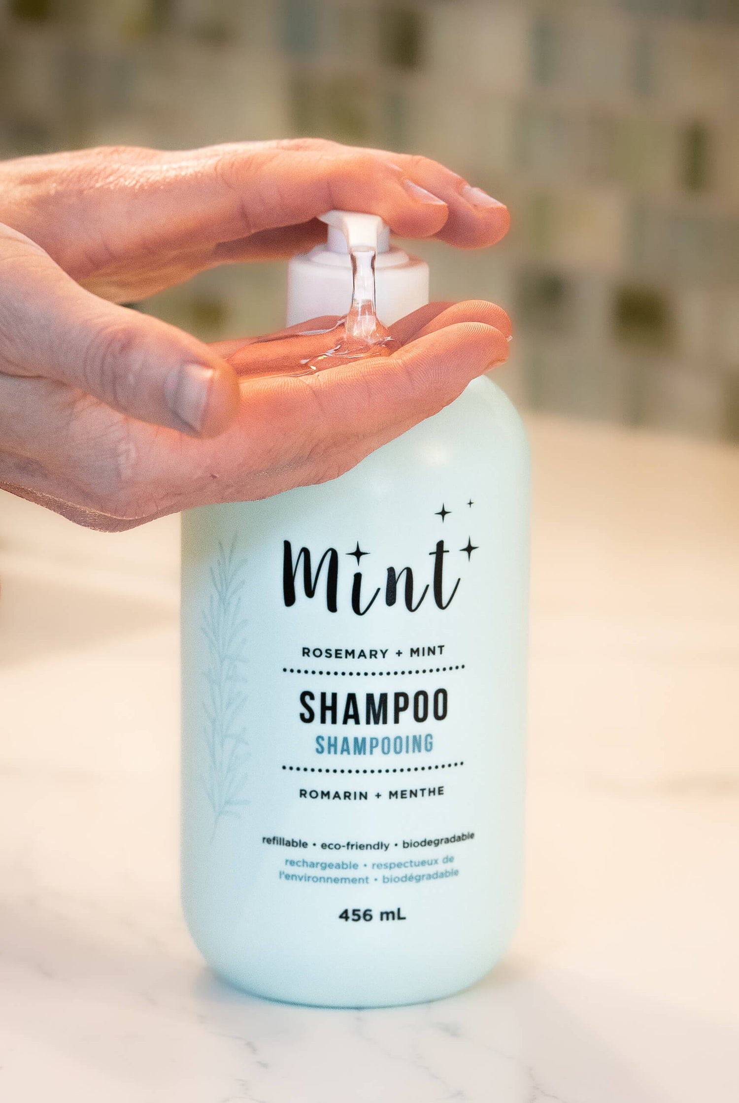 A close-up of a hand dispensing clear liquid shampoo from a light mint-green bottle labeled "Mint Rosemary + Mint Shampoo." The bottle is placed on a white surface with a blurred background.