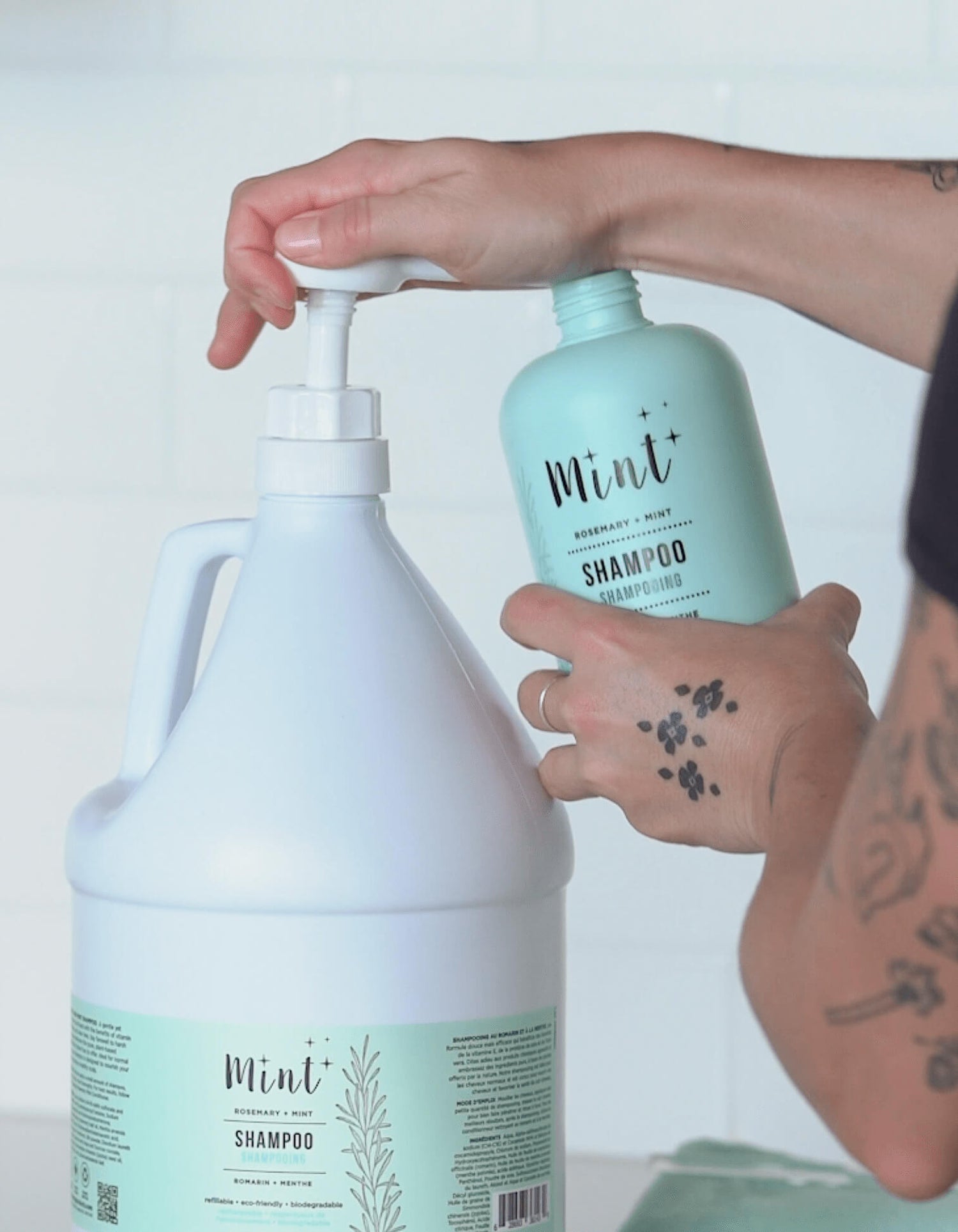 A hand refilling a smaller light mint-green shampoo bottle from a large white refill jug labeled "Mint Rosemary + Mint Shampoo." The background is a white tiled wall.