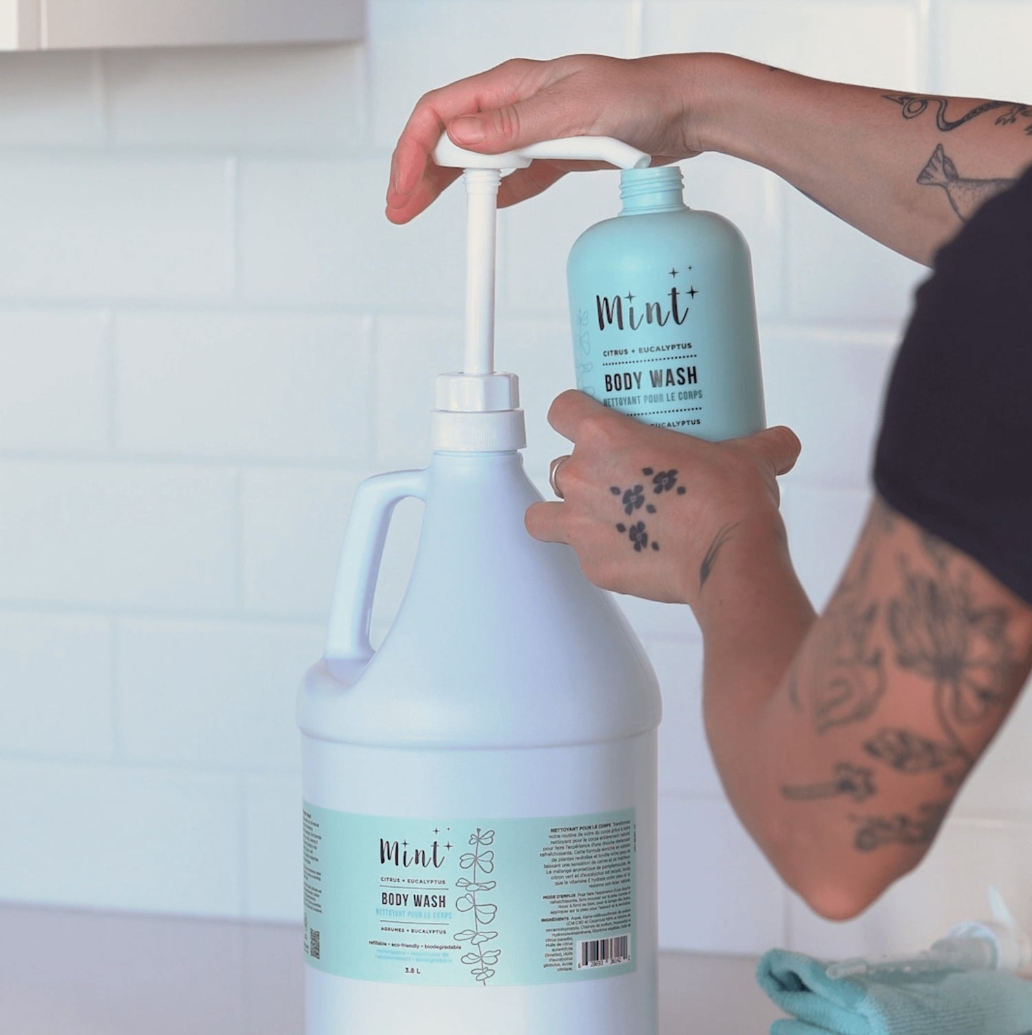 A person refilling a Mint body wash bottle from a large Mint body wash refill jug using a pump dispenser, demonstrating a convenient and eco-friendly refill process.