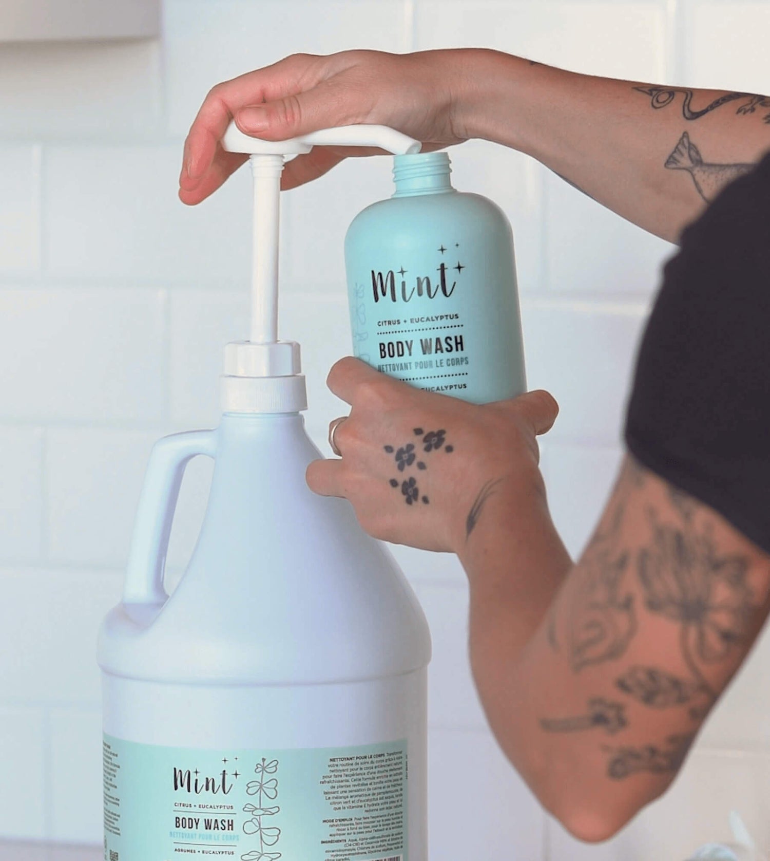 A close-up of a hand refilling a light mint-green "Mint Citrus + Eucalyptus Body Wash" bottle from a large white refill jug. The background features white