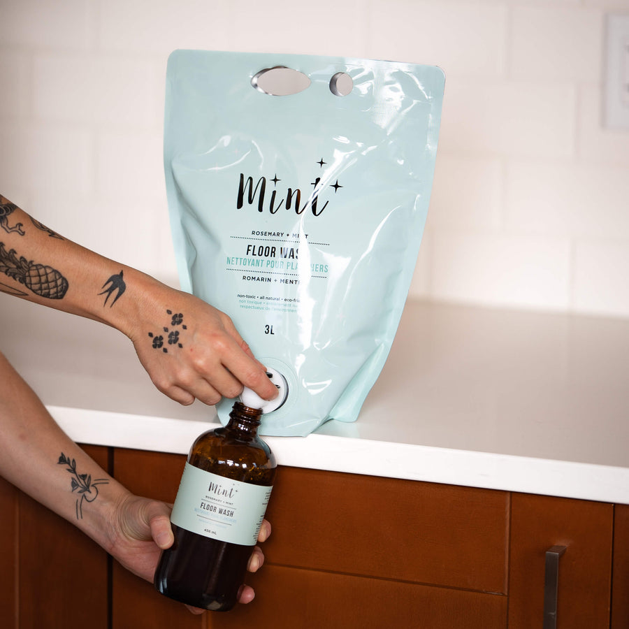 Hand with tattoos refilling a reusable amber glass bottle labeled 'Mint Floor Wash' from a 3L light blue refill pouch of Mint Cleaning Products' floor wash, placed on a white countertop with a wooden cabinet below.