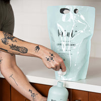 Person with tattooed arms refilling a reusable light blue bottle labeled 'Mint Laundry Detergent' from a 3L light blue refill pouch of Mint Cleaning Products' lavender and mint-scented laundry detergent, placed on a white countertop with a wooden cabinet below.