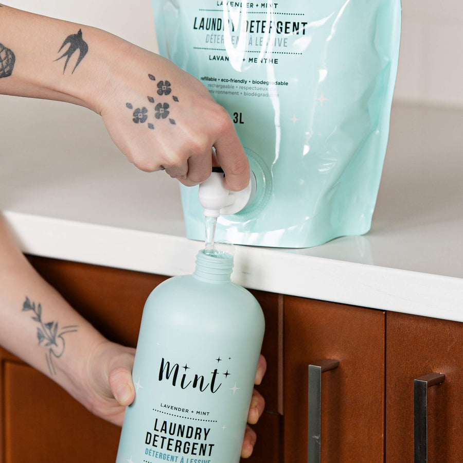 Person with tattooed arms refilling a reusable light blue bottle labeled 'Mint Laundry Detergent' from a 3L light blue refill pouch of Mint Cleaning Products' lavender and mint-scented laundry detergent, placed on a white countertop with a wooden cabinet below.