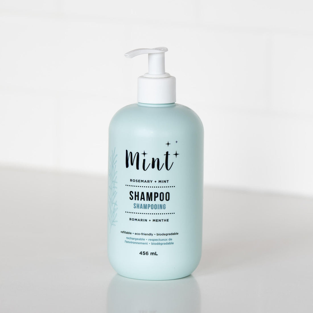 A 456 mL light blue bottle of "Mint+ Shampoo" with a pump dispenser, displayed on a white countertop. The label highlights the scent "Rosemary + Mint" and includes bilingual text, "Shampooing" and "Romarin + Menthe." The product&