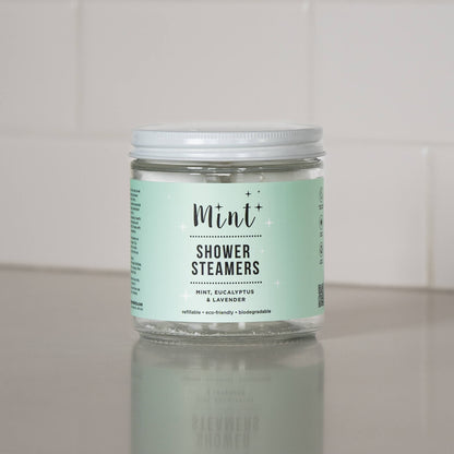 A sealed jar of &quot;Mint+ Shower Steamers&quot; on a sleek, light-colored countertop. The mint green label highlights the scent combination of mint, eucalyptus, and lavender, as well as its eco-friendly features: refillable, biodegradable, and sustainable. The background includes a clean, neutral wall, creating a minimalist and fresh aesthetic.