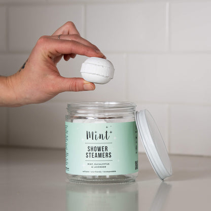 A jar of &quot;Mint+ Shower Steamers&quot; with a mint green label on a white countertop. The label reads &quot;Mint, Eucalyptus &amp; Lavender&quot; and highlights that the product is refillable, eco-friendly, and biodegradable. A hand is holding a single round shower steamer above the open jar, with the lid placed to the side. The background features a clean, white subway tile backsplash.