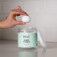 A jar of "Mint+ Shower Steamers" with a mint green label on a white countertop. The label reads "Mint, Eucalyptus & Lavender" and highlights that the product is refillable, eco-friendly, and biodegradable. A hand is holding a single round shower steamer above the open jar, with the lid placed to the side. The background features a clean, white subway tile backsplash.