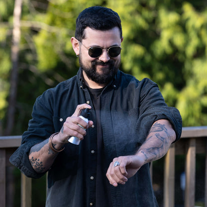 A man wearing sunglasses and a black shirt sprays Mint Outdoor Mist onto his arm while standing outdoors on a deck. The forest is visible in the background.