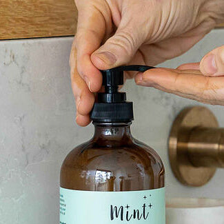 A person pressing a black pump dispenser on a Mint bottle, dispensing liquid soap in a modern bathroom setting