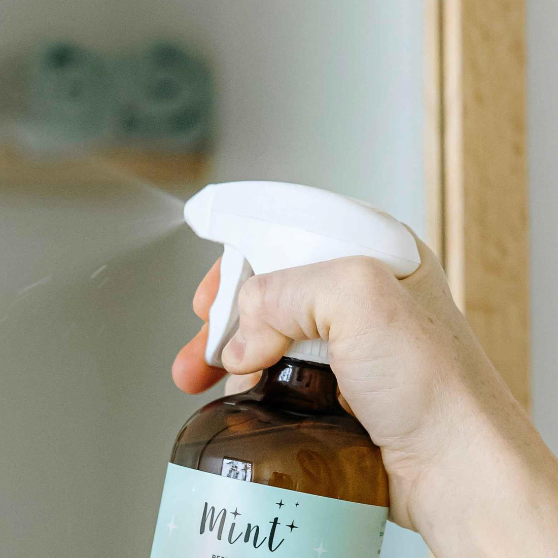 A person using a Mint cleaning bottle with a white trigger sprayer, spraying a fine mist, demonstrating its functionality.